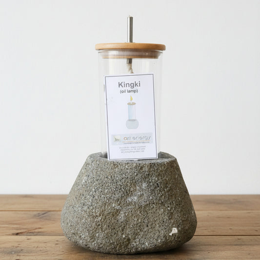 A Kingki oil lamp by art energy, featuring a textured gray stone base, a clear glass jar with a light wood lid, and a white paper label in the center, displayed on a wooden table in a store setting.